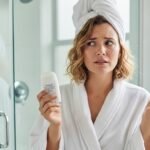Γυναίκα με λευκή ρόμπα κοιτάζει προβληματισμένη ένα αποσμητικό μέσα σε μπάνιο.
