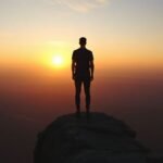 Ένας άνθρωπος κοιτάζει το ηλιοβασίλεμα από ένα βράχο, συμβολίζοντας την ανασκόπηση και την ελπίδα για τη νέα χρονιά.