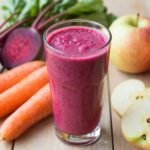 Ένα πολύχρωμο smoothie με παντζάρι, καρότο και μήλο σε ένα ποτήρι, με φρέσκα υλικά γύρω του.