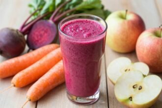 Ένα πολύχρωμο smoothie με παντζάρι, καρότο και μήλο σε ένα ποτήρι, με φρέσκα υλικά γύρω του.