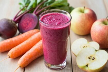 Ένα πολύχρωμο smoothie με παντζάρι, καρότο και μήλο σε ένα ποτήρι, με φρέσκα υλικά γύρω του.