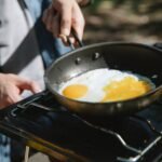 From above of crop faceless male cooker frying eggs on metas gas burner using skillet in nature