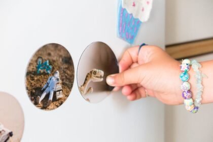 A child's hand with bracelets holding a dinosaur magnet on a refrigerator adorned with stickers.