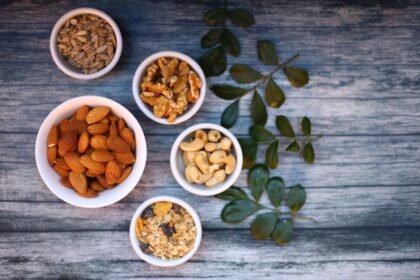 Aerial view of assorted nuts and seeds in white bowls with green leaves on a rustic wooden table.