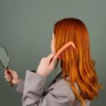 Red-haired woman using a comb while holding a mirror, focusing on hair care and beauty.