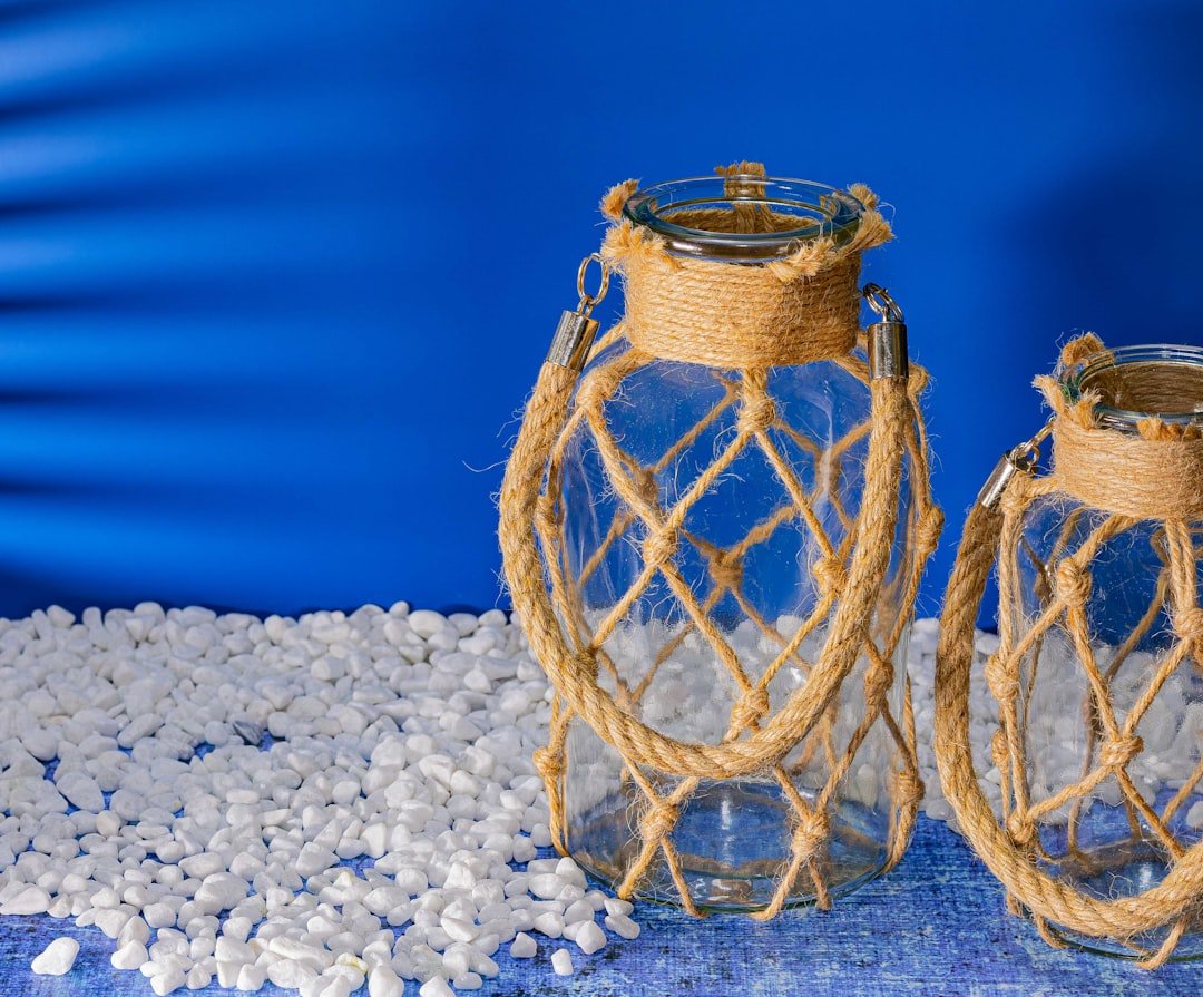 Two glass lanterns with rope accents on white pebbles.
