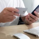 Person holding credit card and smartphone for an online transaction at a desk.